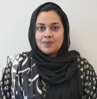 Close-up head-and-shoulders portrait of a person wearing a dark patterned headscarf, facing forward against a plain light-colored background.